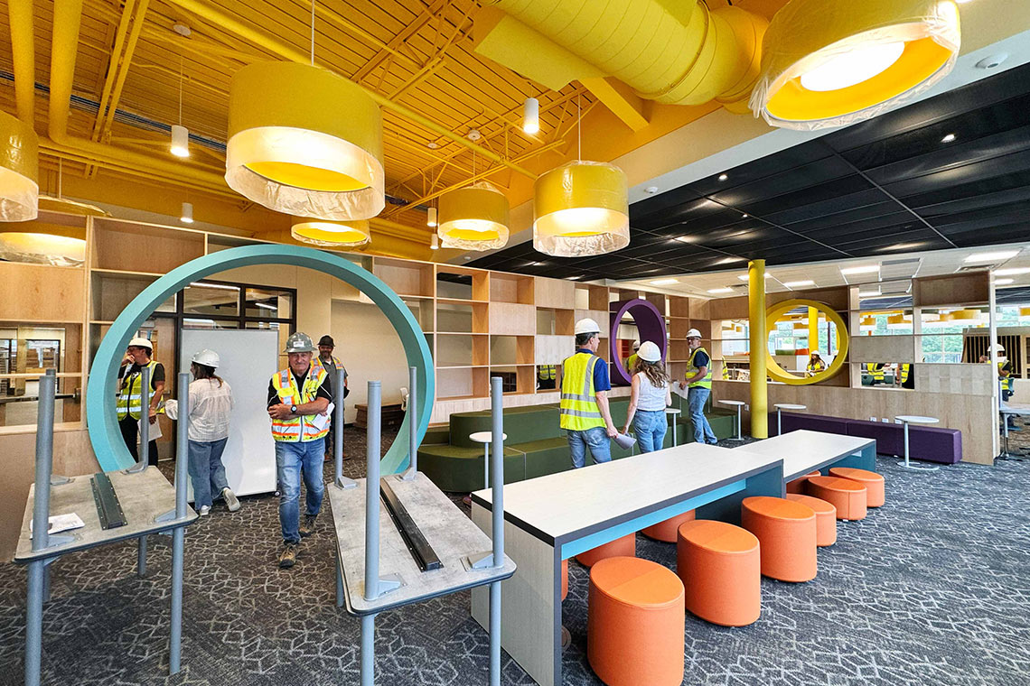 An elementary media center with bright yellow ceilings, a circular doorway, long tables and chairs.